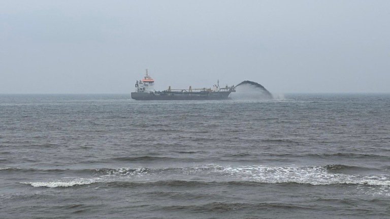 Een schip voert baggerwerkzaamheden uit op zee, waarbij een grote boog van materiaal in het water wordt gespoten, omgeven door een bewolkte lucht en kalme golven.